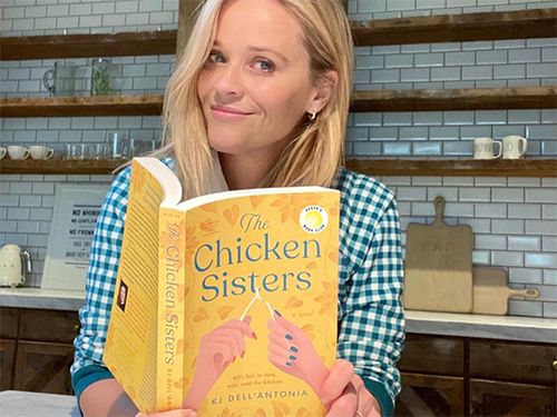 Reese Witherspoon holding a book titled The Chicken Sisters in a kitchen.