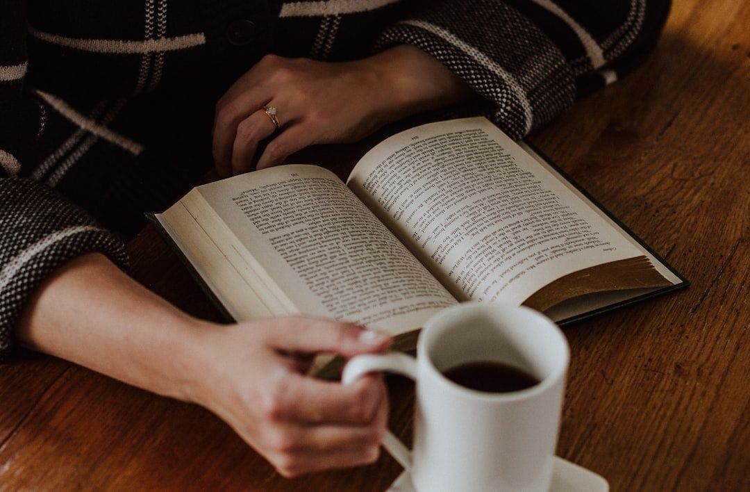 Person reading an open book beside a cup of coffee at a wooden table